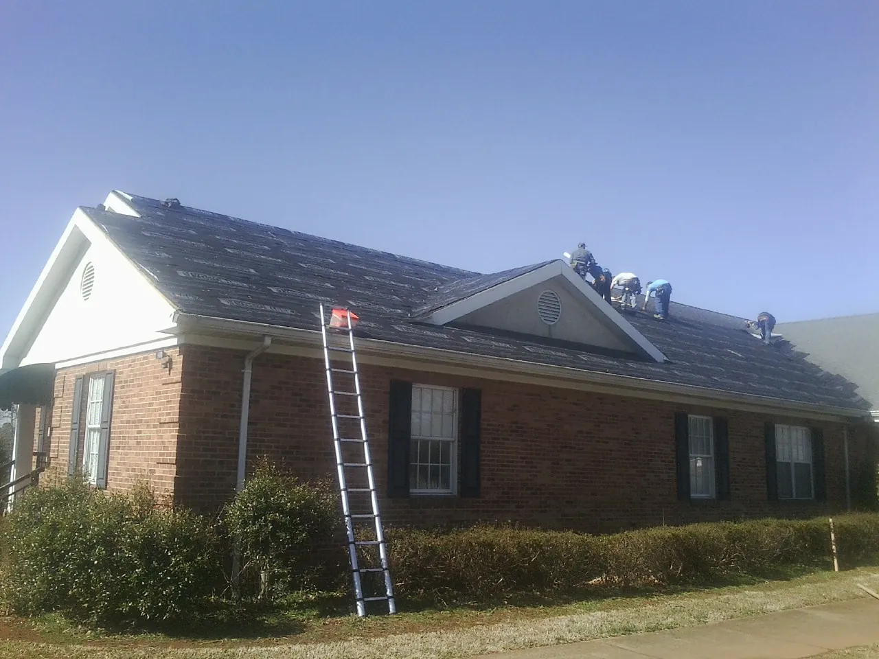 Crew tearing shingles off a brick ranch roof with a ladder set — Metal Roof Repair work by our crew in Burnettsville