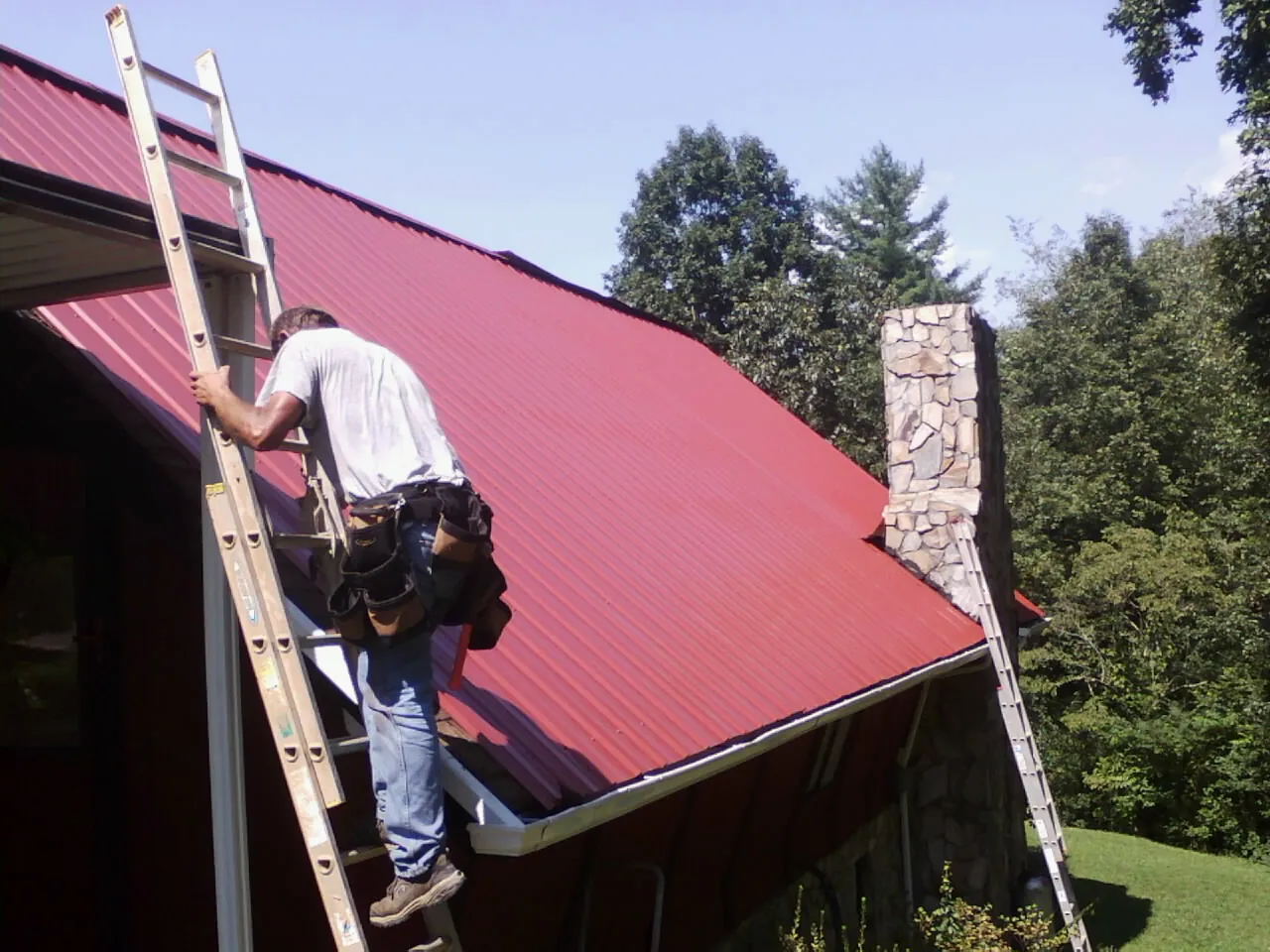 Roofer on a ladder beside a red metal roof and stone chimney — Soffit & Fascia Repair by Roofing Brothers in Burnettsville